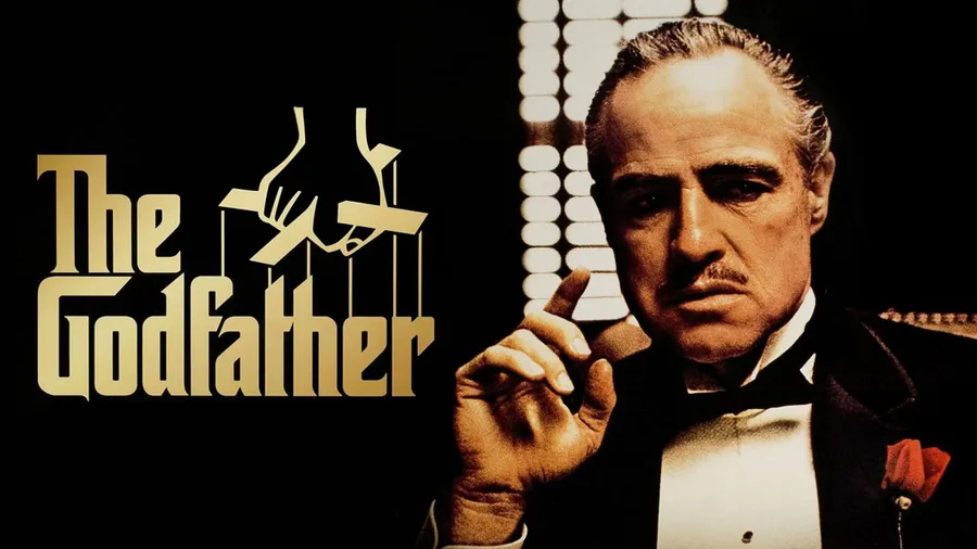 A dark, dramatic scene from the movie 'The Godfather,' featuring a powerful, shadowy figure of Don Vito Corleone seated in a dimly lit room. He is dressed in a black tuxedo with a red rose pinned to his lapel, embodying authority and control. His face, partially obscured by shadows, displays a stern and contemplative expression, capturing the tension and gravity of his role as the head of a powerful mafia family. The atmosphere is filled with sober tones, highlighting the film's themes of power, loyalty, and betrayal. Top Oscar-Winning Movie You Must Watch