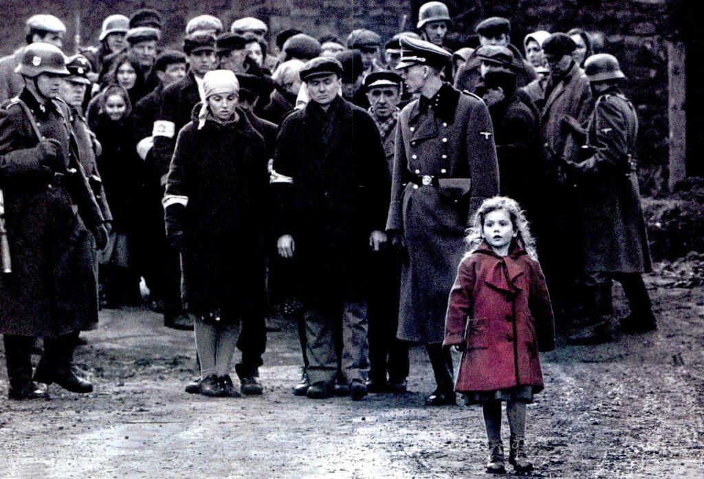 A striking scene from the film 'Schindler's List,' depicting a group of Jewish prisoners being watched by Nazi soldiers. The image is predominantly in black and white, except for a young girl in the foreground wearing a bright red coat, who stands out amidst the somber tones. The girl appears to be walking alone, while the prisoners in the background are huddled together under the cold, watchful gaze of armed soldiers. The contrast between the girl’s red coat and the surrounding grayscale emphasizes her innocence and the grim realities of the Holocaus. 
One of the Best Oscar-Winning Films Of All Time